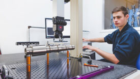 Coordinate measuring machine (CMM) . The device is being programmed by a young male to measure the physical geometrical characteristics of an object. The operator is concentrating on the procedure. A display screen is seen in the background.
