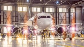 Large-scale inspection of all aircraft systems in the aircraft hangar by worker mechanics and other specialists. Bright light streaming outside from the garage door.