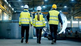 Shot of Three Engineers Walking Through a Heavy Industry Manufacturing Factory. In the Background Welding Work in Progress, Various Metalwork, Pipeline/ Barrel Components.