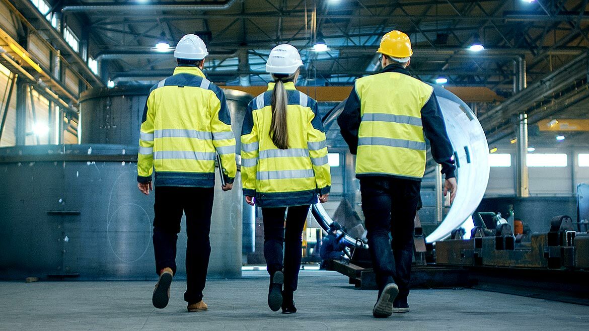Shot of Three Engineers Walking Through a Heavy Industry Manufacturing Factory. In the Background Welding Work in Progress, Various Metalwork, Pipeline/ Barrel Components.