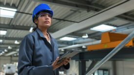 Woman engineer in a blue helmet and work coat posing at a factory holding a tablet.