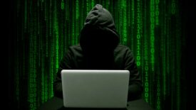 Hacker in a dark hoodie committing cyber crime with a laptop, green binary numbers on a black background.