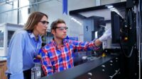Business colleagues wearing safety glasses and using computer in industrial production line.