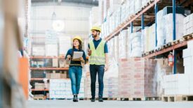 Two employees in hardhats checking inventory on warehouse racks.