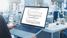 An engineer holds a laptop with mock-up charts. In the blurred background, three people are working together at a desk.