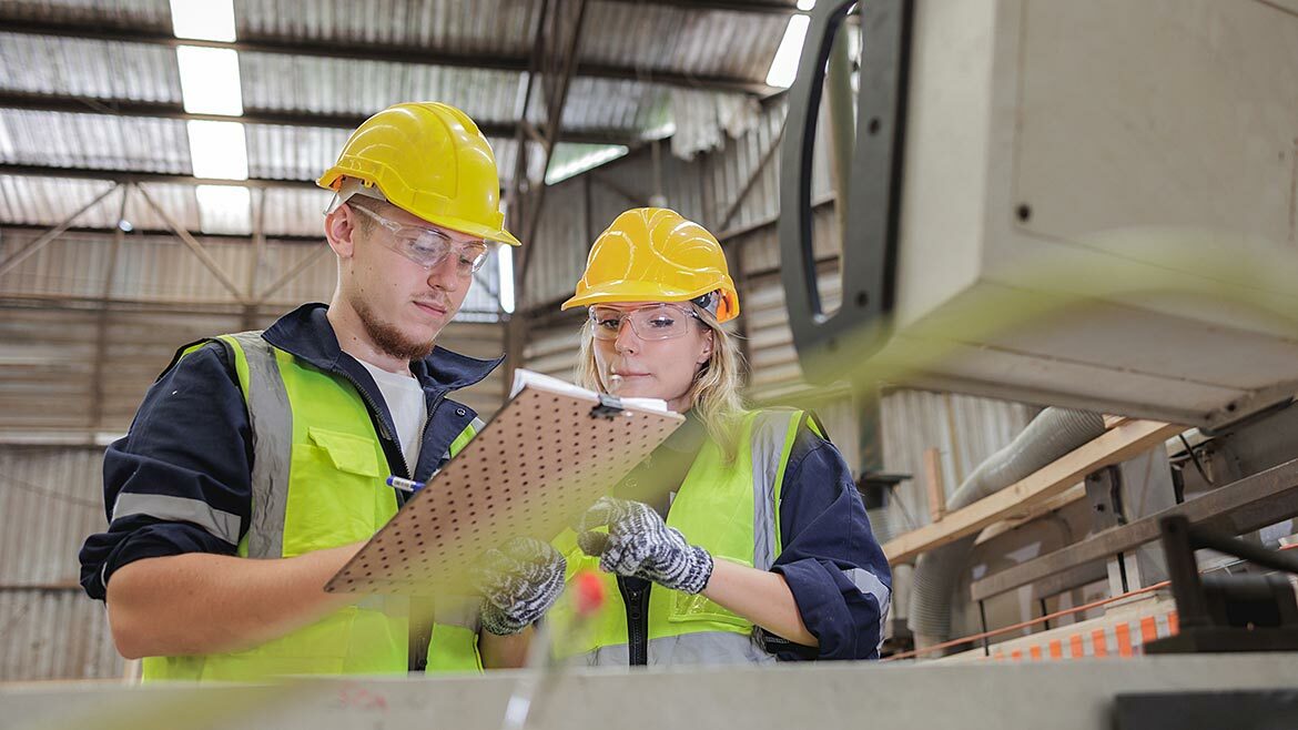A young male and young female engineer in hardhats and safety vests, looking at a clipboard in a warehouse.