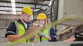A young male and young female engineer in hardhats and safety vests, looking at a clipboard in a warehouse.